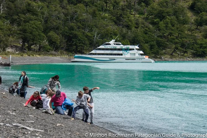 Personas exploran una orilla rocosa con un barco de excursión amarrado al fondo.